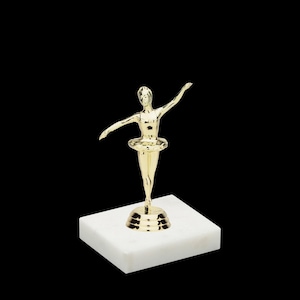 May include: A gold-colored ballerina figurine on a white marble base. The ballerina is wearing a tutu and is posed in a graceful dance position.