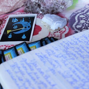 May include: A tarot card with a blue crescent moon and a yellow stick figure holding a broom. The card is surrounded by other tarot cards and crystals on a patterned tablecloth.