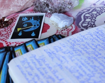 May include: A tarot card with a blue crescent moon and a yellow stick figure holding a broom. The card is surrounded by other tarot cards and crystals on a patterned tablecloth.