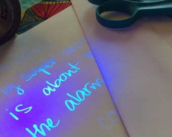 May include: A close-up of a white piece of paper with blue ink that glows under a black light. The writing says "John is super secret about the alarm".