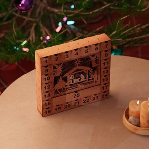 Light wooden nativity advent calendar placed on a round table near lit candles and a decorated Christmas tree.
