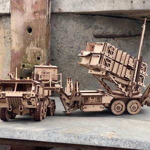 May include: A wooden model kit of a military missile launcher truck featuring a detailed cab, multi-axle trailer, and an adjustable launch platform with rectangular storage containers. The construction displays intricate laser-cut wood panels.
