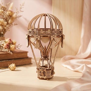 Op de afbeelding: Een gedetailleerd model van een luchtballon van licht hout. Het model heeft een mand, decoratieve linten en een gedetailleerde bovenstructuur. De ballon staat op een licht oppervlak en is een decoratief item.
