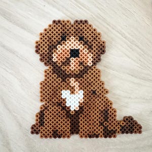 Puede incluir: Un perro marrón y canela hecho de cuentas de plástico fusionadas. El perro tiene una mancha blanca en el pecho y una nariz negra. El perro está sentado con la cola enrollada. Las cuentas están fusionadas para crear un efecto pixelado.