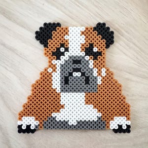 Op de afbeelding: Handgemaakte hondenfiguur gemaakt van kleurrijke plastic kralen. De hond heeft een bruin en wit lichaam, een grijze snuit en zwarte oren. Het gepixelde ontwerp geeft het een unieke, speelse uitstraling.