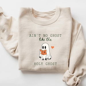 May include: Beige sweatshirt with the text "Ain't no ghost like the Holy Ghost" in green and black. A cartoon ghost holding a book with an orange cover and a small orange heart are also featured on the sweatshirt.