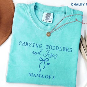 Può includere: Una maglietta turchese con le parole "CHASING TODDLERS and Jesus MAMA OF 3" stampate in blu. Sono visibili anche una collana con pendente dorato e un cappello marrone. La maglietta è etichettata "CHALKY MINT".