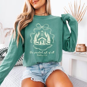 Puede incluir: Sudadera de color verde azulado con un gráfico crema de una escena de la natividad, árboles y una estrella. El texto "the greatest gift of all" y "Matthew 1:21" están impresos debajo. La sudadera tiene mangas largas.