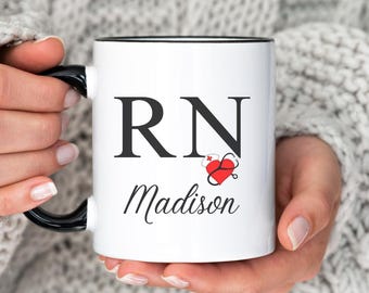 May include: White ceramic mug with a black rim and handle. The mug has a black and red graphic of a stethoscope and heart with the letters "RN" and the name "Madison" written in black cursive.
