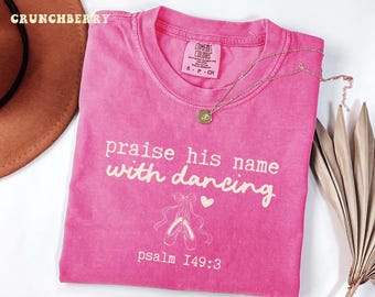 May include: A bright pink t-shirt with the words "praise his name with dancing" and the Bible verse Psalm 149:3 printed in white. The shirt features a ballet shoe graphic and a gold necklace.