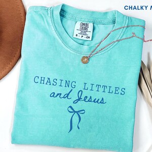 Può includere: Una maglietta turchese con la scritta "CHASING LITTLES and Jesus" in blu. Una grafica a fiocco blu è sotto il testo. Una collana con pendente dorato e un cappello marrone sono anch'essi nell'immagine. La maglietta è etichettata "Chalky Mint".