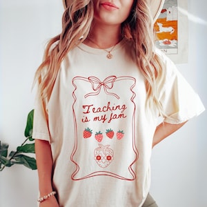 May include: Cream-colored t-shirt with a red graphic of a jam jar, strawberries, and the text "Teaching is my jam." The design includes a bow at the top and a smiling strawberry face at the bottom. The shirt has short sleeves.