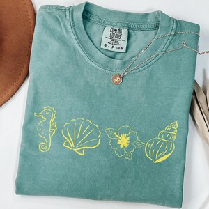 Peut inclure: Un t-shirt Comfort Colors vert d'eau avec des graphiques jaunes sur le thème de la mer, dont un hippocampe, un coquillage, une fleur et un coquillage. Un collier argenté avec un pendentif repose sur le t-shirt. Un chapeau marron et une feuille de palmier séchée sont en arrière-plan.