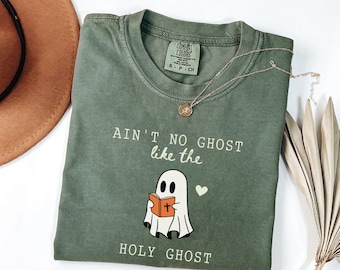 Op de afbeelding: Olijfgroen t-shirt met de tekst "Ain't no ghost like the Holy Ghost" en een cartoon spook dat een boek leest. Een gouden ketting en een gedroogde plant zijn ook zichtbaar.