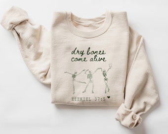 May include: A cream-colored sweatshirt with the text "dry bones come alive" in green script. Below the text are three dancing skeleton figures. The sweatshirt also includes the Bible verse Ezekiel 37:5.