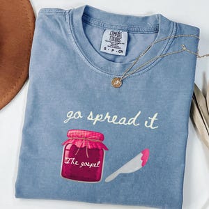 以下が含まれることがあります： 「go spread it」の文字と、ジャムの瓶とナイフのグラフィックが描かれた水色のTシャツ。「The gospel」の文字が瓶の下にあります。金のネックレスがシャツの上にかけられています。