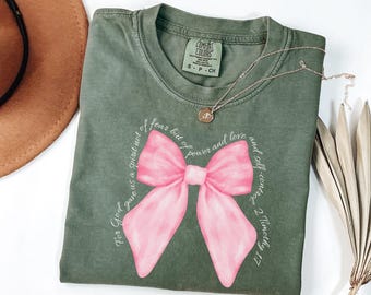 May include: An olive green t-shirt featuring a pink bow design and the text "For God gave us a spirit not of fear but of power and love and self-control 2 Timothy 1:7". A gold necklace with a pendant is on the shirt.