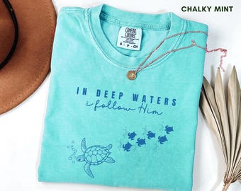 May include: A chalky mint green t-shirt with the text "IN DEEP WATERS I follow Him" and a turtle graphic. The shirt has a crew neck and short sleeves. A gold pendant necklace is draped across the shirt.