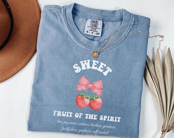May include: A light blue t-shirt with a white graphic of two red cherries with a red and white gingham bow. The text "SWEET FRUIT OF THE SPIRIT" is printed below the cherries, with the words "love joy peace patience kindness goodness faithfulness gentleness self-control" printed below the text.