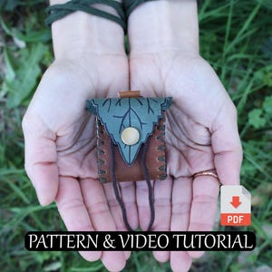 May include: A small, handcrafted leather pouch with a leaf-shaped flap in shades of green and brown, secured with a button. The text "PATTERN & VIDEO TUTORIAL" is displayed at the bottom. The pouch is held in open hands against a backdrop of green foliage.