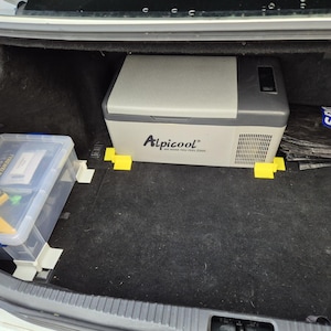 May include: A grey portable refrigerator with the text "Alpicool" on the front, sitting in the back of a car. The refrigerator is on a black carpeted surface and is surrounded by yellow and blue plastic blocks.
