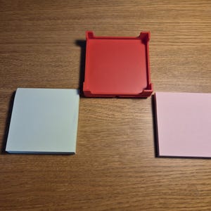 May include: A red square tray with raised edges sits above two stacks of square sticky notes. One stack is light blue, the other is pink. The items are arranged on a wooden surface.