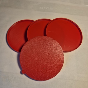 May include: Four circular red coasters with a textured top surface and a smooth raised rim. The coasters are arranged in a cluster, with one piece positioned in the foreground and three pieces partially overlapping behind it, featuring a small tab.