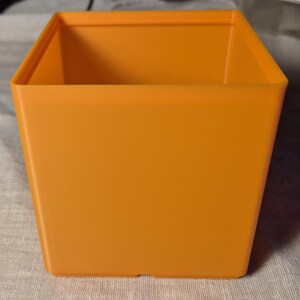 May include: An orange square plastic plant pot with a slightly tapered design. The pot has a smooth, matte finish and is empty. The container is suitable for indoor or outdoor use.