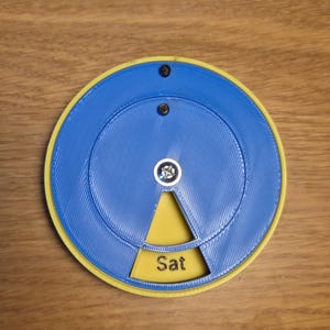 May include: A circular, 3D-printed object with a blue outer ring and a yellow inner section. The centre features a silver-coloured component and the word "Sat" in white. Two black screws secure the top.