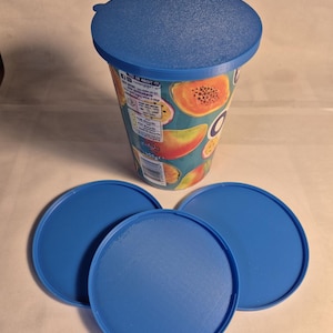 May include: A cylindrical food container with a tropical fruit print featuring papayas and mangoes, topped with a blue plastic lid. Three additional matching blue plastic lids are arranged in a semi-circle in front of the container.