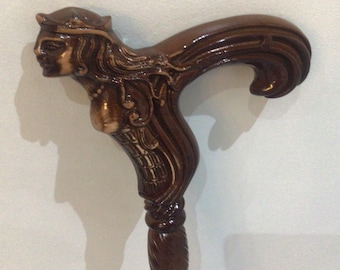 May include: A dark brown wooden walking stick with a carved handle depicting a woman's face and flowing hair. The handle is intricately carved and curved for grip. The shaft of the cane has a spiral design.