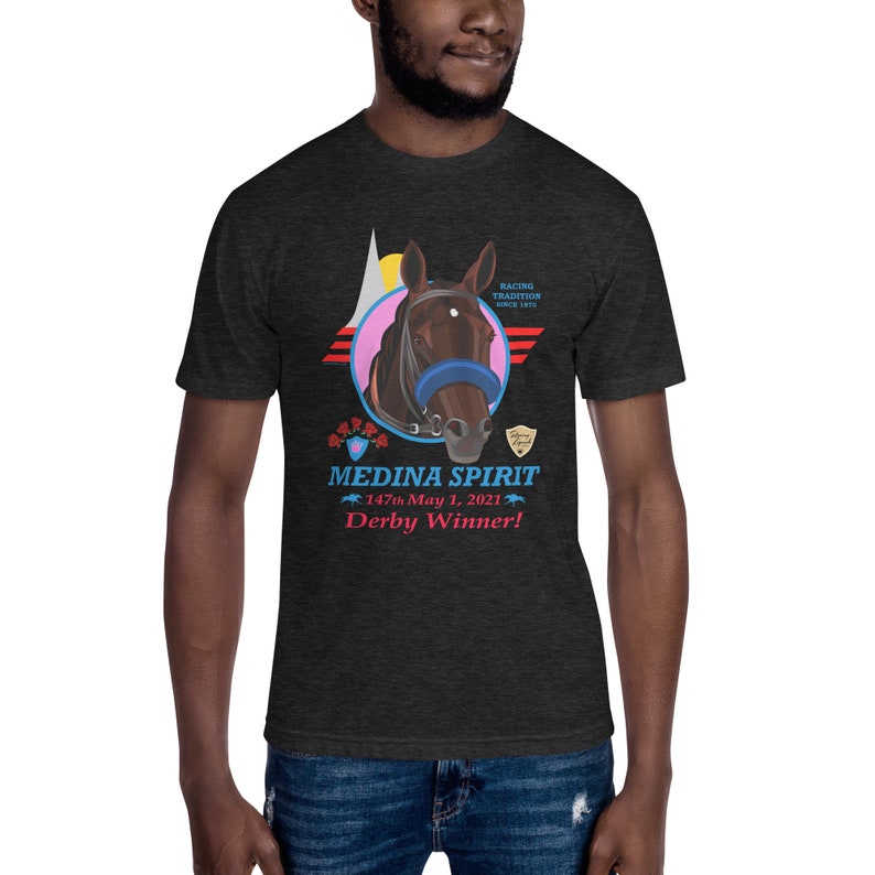 Puede incluir: Camiseta gris oscuro con un gr&aacute;fico de cabeza de caballo y el texto "MEDINA SPIRIT Derby Winner!" El dise&ntilde;o incluye un c&iacute;rculo rosa, detalles en rojo y azul y las palabras "Racing Tradition Since 1875."