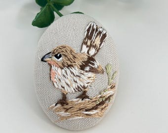 May include: A brown and white embroidered bird perched on a branch, stitched on a beige oval fabric. The bird has a detailed design with brown, white, and beige thread.