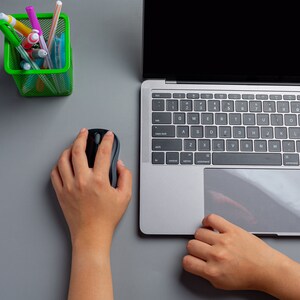 Könnte beinhalten: Die Hände einer Person, die eine schwarze drahtlose Computermaus auf einer grauen Oberfläche benutzen. Ein Laptop mit schwarzem Bildschirm ist auf der Oberfläche geöffnet. Ein grüner Drahtkorb mit bunten Stiften und Bleistiften befindet sich in der oberen linken Ecke des Bildes.