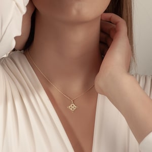 Pu&ograve; includere: Una collana color oro con un ciondolo a forma di quadrifoglio. Il ciondolo presenta un motivo dettagliato con piccoli accenti scuri. La collana &egrave; indossata su una camicetta bianca.