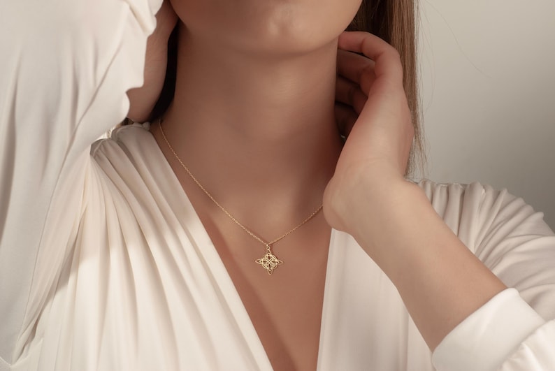 Pu&ograve; includere: Una collana color oro con un ciondolo a forma di quadrifoglio. Il ciondolo presenta un motivo dettagliato con piccoli accenti scuri. La collana &egrave; indossata su una camicetta bianca.