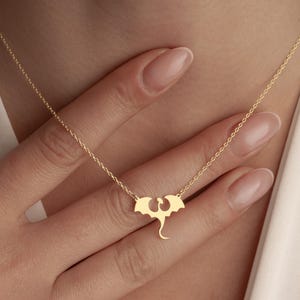 May include: A gold-colored necklace featuring a dragon pendant. The dragon has outstretched wings and a curved tail. The necklace is on a delicate chain, and the pendant is centered. The background is a soft, neutral color.