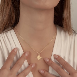 Pu&ograve; includere: Una collana color oro con un ciondolo a forma di stella a quattro punte. La collana &egrave; indossata da una persona che indossa un top bianco. Il ciondolo ha un design traforato. La catena della collana &egrave; delicata.
