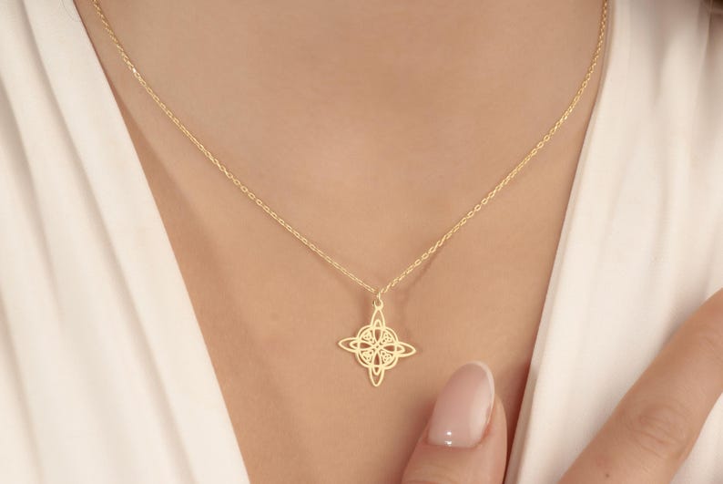 Pu&ograve; includere: Una delicata collana color oro con un ciondolo dal design traforato a quattro punte. Il ciondolo pende da una sottile catena, appoggiato sul petto di una persona. Lo sfondo &egrave; un capo bianco.