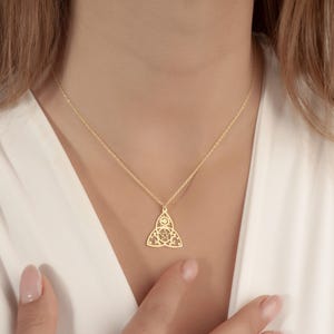 May include: A gold-colored pendant necklace featuring a Celtic knot design. The pendant is suspended from a delicate chain. The necklace is worn against a white top.