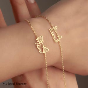 Puede incluir: Dos pulseras personalizadas de color dorado. Cada pulsera presenta un dije en forma de gato con un nombre grabado. Las pulseras están conectadas por delicadas cadenas. Las pulseras se usan en la muñeca.