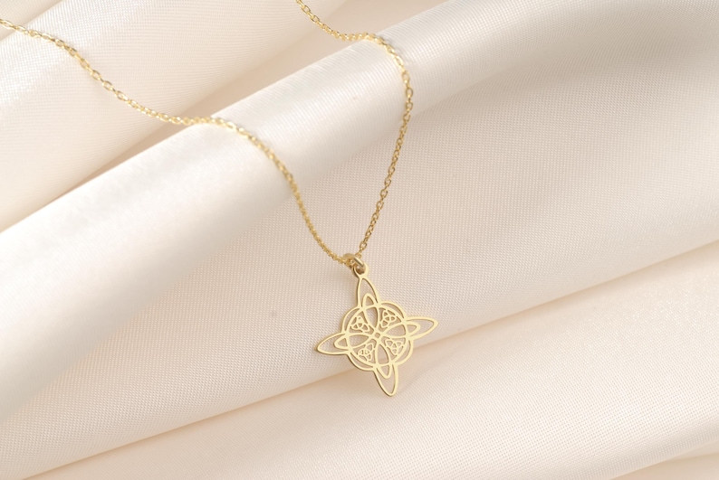 Pu&ograve; includere: Una collana con pendente dorato con un design a nodo a quattro punte. Il pendente &egrave; appeso a una delicata catena, su uno sfondo di morbido tessuto bianco sporco. La collana &egrave; un gioiello.