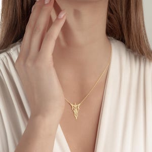 May include: A gold-colored necklace with a pendant featuring an intricate, symmetrical design. The necklace hangs on a delicate chain. The person is wearing a white top.