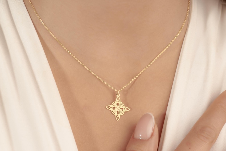 Pu&ograve; includere: Una delicata collana color oro con un ciondolo dal design traforato a quattro punte. Il ciondolo pende da una sottile catena, appoggiato sul petto di una persona. Lo sfondo &egrave; un capo bianco.