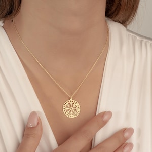 May include: A gold-colored pendant necklace featuring a circular pendant with an intricate design. The pendant hangs from a delicate chain. The necklace is worn against a white blouse.