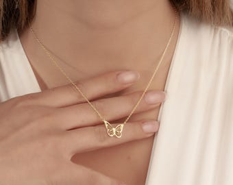 Pode incluir: Um delicado colar com pingente de borboleta dourada. O design da borboleta é intrincadamente detalhado. O colar é usado por uma pessoa com uma parte superior branca. A corrente é fina e elegante.