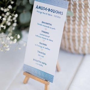 May include: A white menu card with blue Greek key border and text, displayed on a small wooden easel. The menu lists appetizers in French, including tomato-mozzarella, salmon-avocado, and feta-honey.