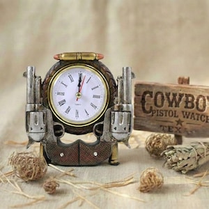 May include: A decorative clock with a western theme. The clock face is surrounded by a bronze-colored frame and two pistol replicas. A small wooden sign reads "COWBOY PISTOL WATCH TEX". The clock hands point to 1:00.
