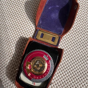 May include: A vintage light meter in a brown leather case. The meter has a silver face with black markings and a red ring around the outer edge. The case has a blue velvet lining.