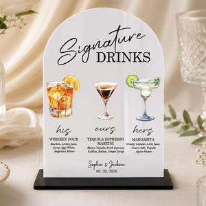 Könnte beinhalten: Ein bogenförmiges weißes Acrylschild mit der Aufschrift Signature Drinks und Illustrationen von Whiskey Sour, Espresso Martini und Margarita. Der Text listet Zutaten, die Namen Sophia & Jackson sowie das Datum 20.08.2026 auf einem schwarzen Sockel.